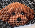May include: A pixel art dog's face created from small, cylindrical beads in shades of brown. The dog has a black nose and white eyes. The artwork is set against a black and white grid, with tweezers in the background.