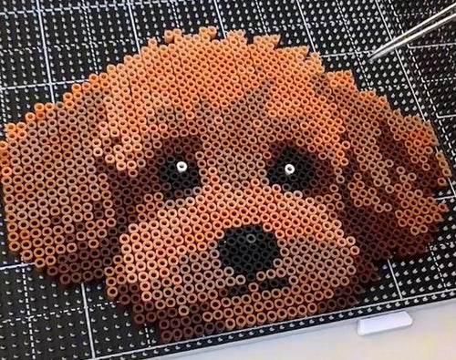 May include: A pixel art dog's face created from small, cylindrical beads in shades of brown. The dog has a black nose and white eyes. The artwork is set against a black and white grid, with tweezers in the background.