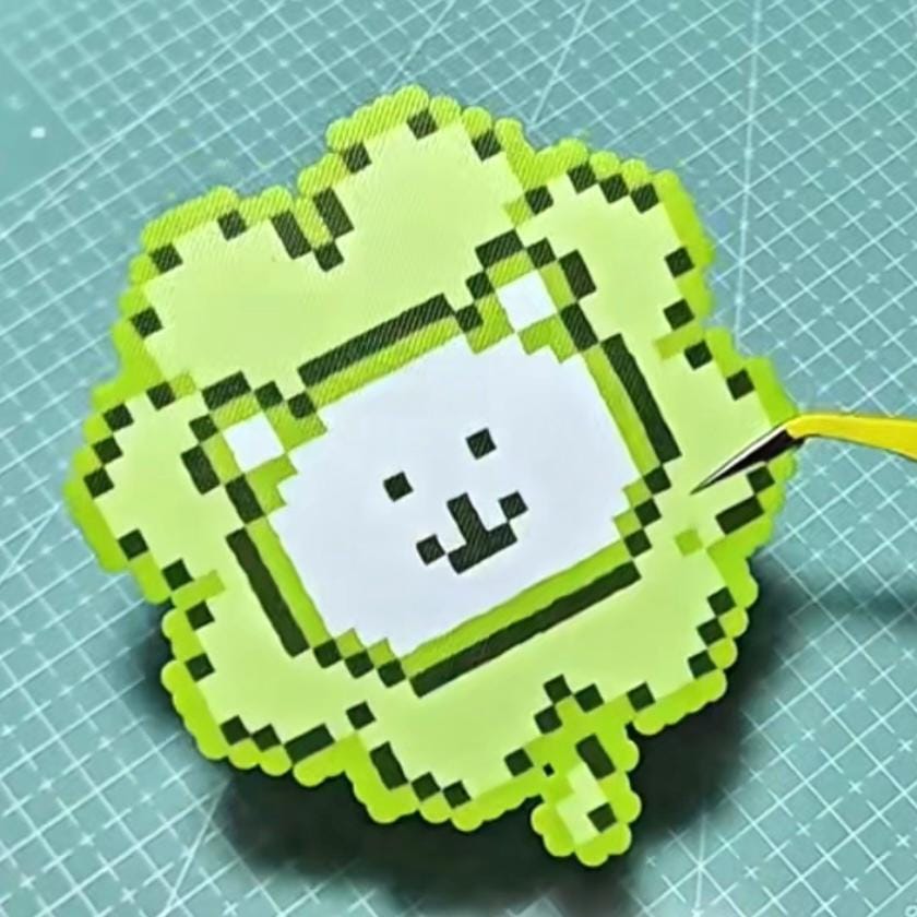 May include: A pixelated green clover-shaped craft with a white square featuring a smiling face. The clover has a dark green outline and a lighter green fill. A pair of yellow tweezers is positioned near the clover.