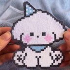May include: A pixelated white puppy-like figure with a blue bow and collar, pink cheeks, and black eyes and nose. The figure is made of small, colourful squares, giving it a retro, handmade appearance. The figure is a cute, whimsical design.