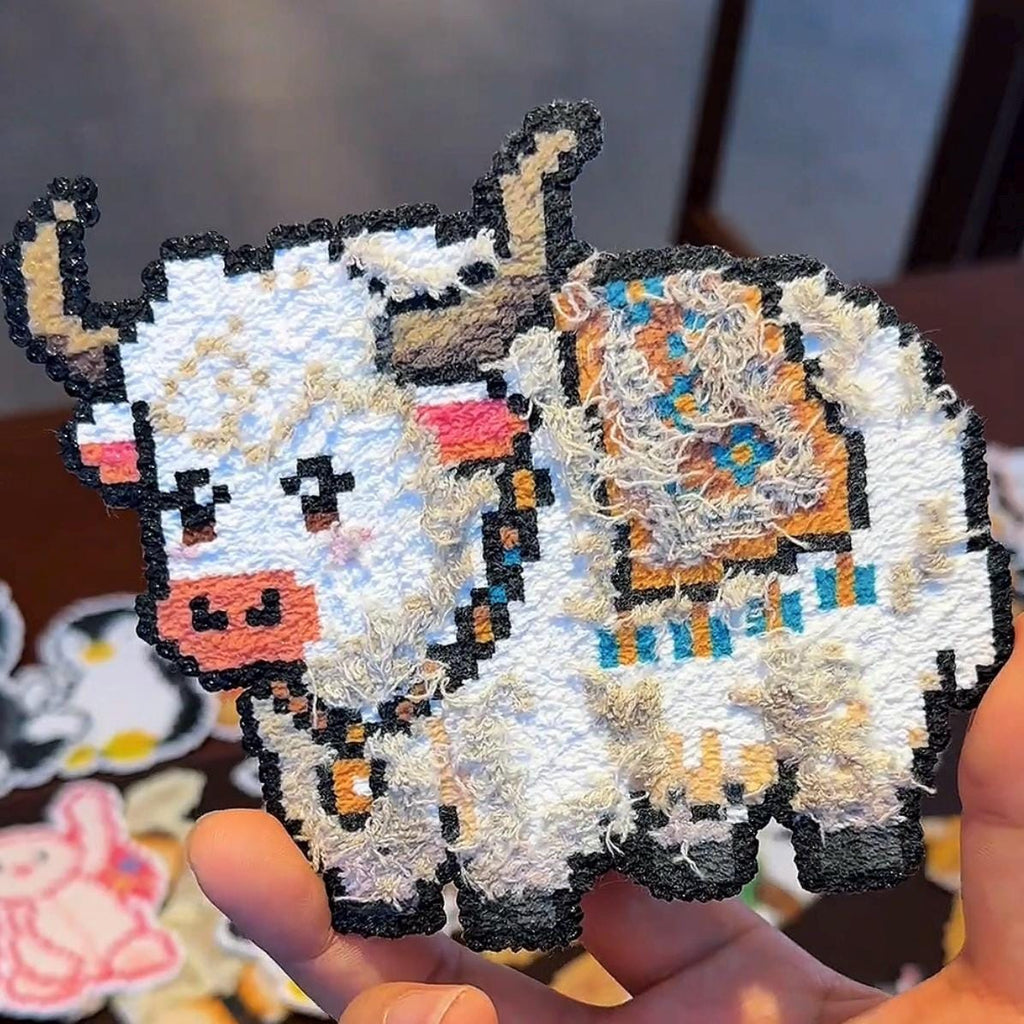 May include: A pixelated yak-shaped patch with a white body, black outline, and textured fur details. The yak has brown horns, a pink muzzle, and colourful accents on its back. The patch is held in a hand.