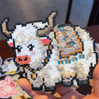 May include: A pixelated yak-shaped patch with a white body, black outline, and textured fur details. The yak has brown horns, a pink muzzle, and colourful accents on its back. The patch is held in a hand.