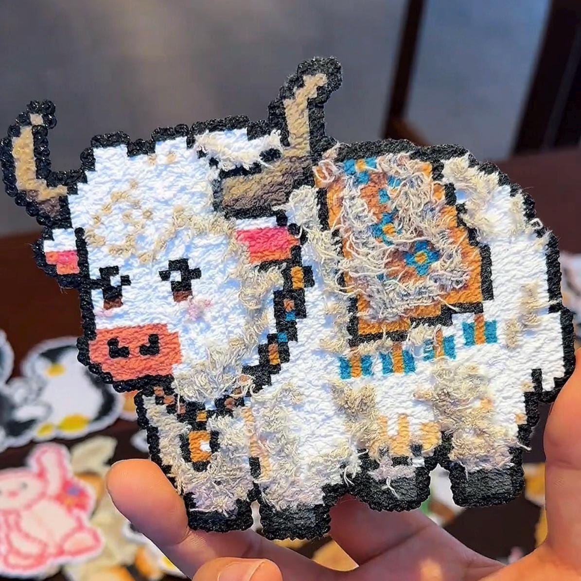 May include: A pixelated yak-shaped patch with a white body, black outline, and textured fur details. The yak has brown horns, a pink muzzle, and colourful accents on its back. The patch is held in a hand.