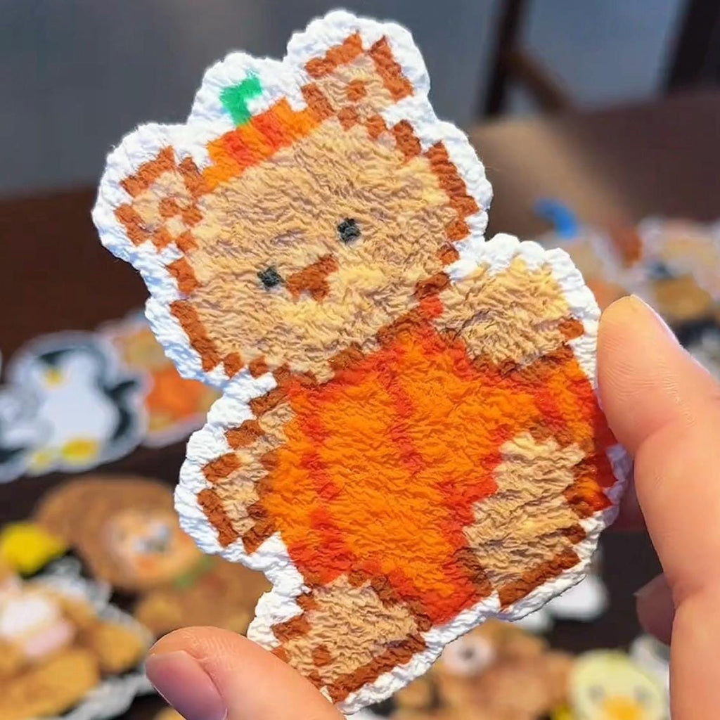 May include: A pixelated teddy bear patch, featuring an orange outfit and a green accent on its head. The bear has a white border and is held in a hand. Other similar patches are visible in the background.