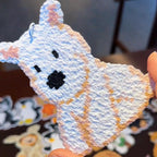 May include: A white, textured cat-shaped keyring with orange and pink accents. The cat has black eyes and a black nose. The keyring is held by a person's fingers. Other keyrings are visible in the background.