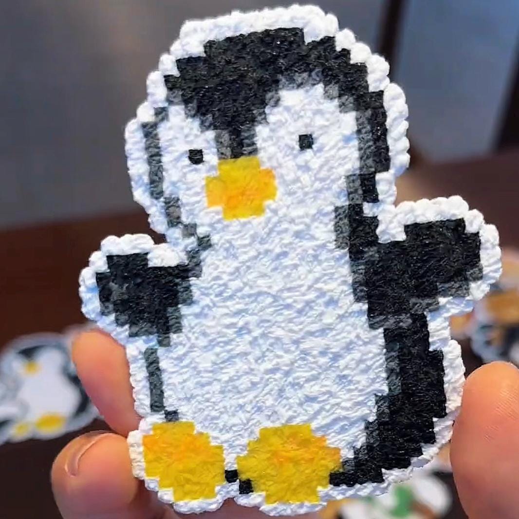 May include: A pixelated penguin-shaped craft with a textured, white body, black head and wings, and yellow feet and beak. The penguin is held up by a hand. The craft has a 3D appearance.