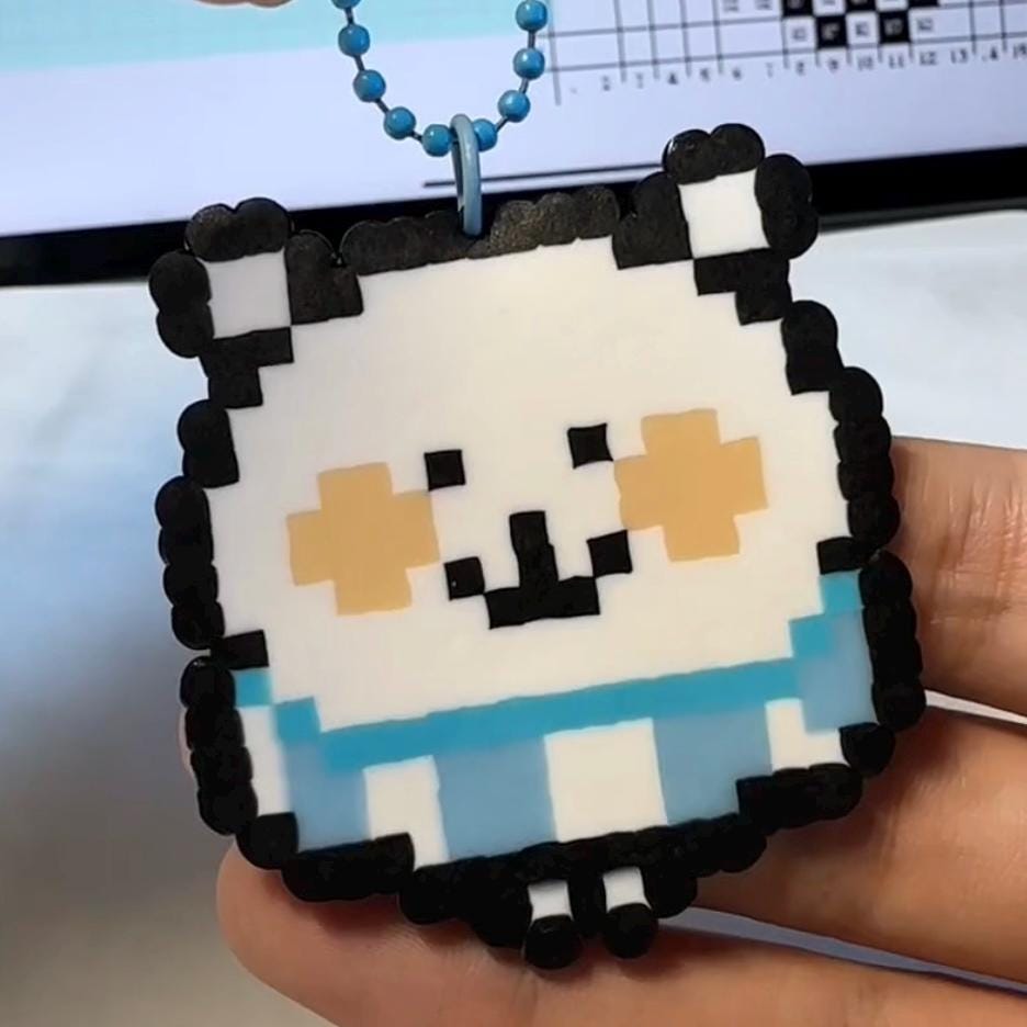 May include: A pixelated white cat charm with black outlines, light blue accents, and peach-coloured cheeks. The charm is attached to a blue beaded chain. The cat has a simple, blocky design, reminiscent of retro video game graphics.