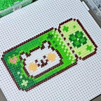 May include: A pixel art creation of a green and white design, possibly a bookmark or a similar item. The design features a cartoon character with a white face, tan cheeks, and a small pink heart. The border is brown.