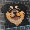 May include: A pixel art depiction of a Rottweiler dog's face, crafted from small black, tan, and brown beads. The dog's features are well-defined, with emphasis on the eyes and snout. The artwork is set against a grid background.