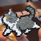 May include: A pixelated cat figurine in grey, white, and black. The cat has a fluffy grey body, white paws, and a black outline. A small yellow detail is visible on the cat's ear. The figurine is held in a person's hand.