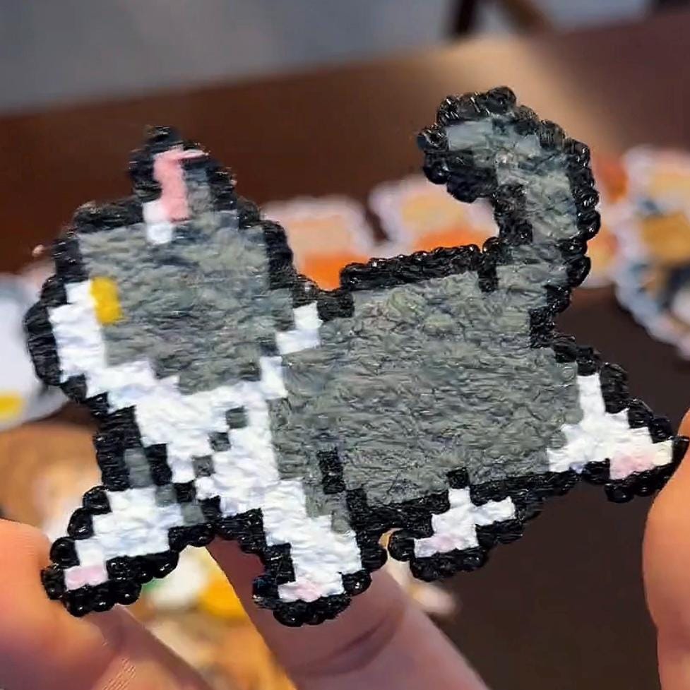 May include: A pixelated cat figurine in grey, white, and black. The cat has a fluffy grey body, white paws, and a black outline. A small yellow detail is visible on the cat's ear. The figurine is held in a person's hand.