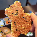 May include: A small, orange teddy bear-shaped charm with a textured surface and black dot details. The charm has a small metal loop at the top for attachment. The bear's face features small black eyes and a stitched-like nose and mouth. The charm is