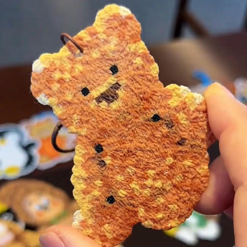 May include: A small, orange teddy bear-shaped charm with a textured surface and black dot details. The charm has a small metal loop at the top for attachment. The bear's face features small black eyes and a stitched-like nose and mouth. The charm is