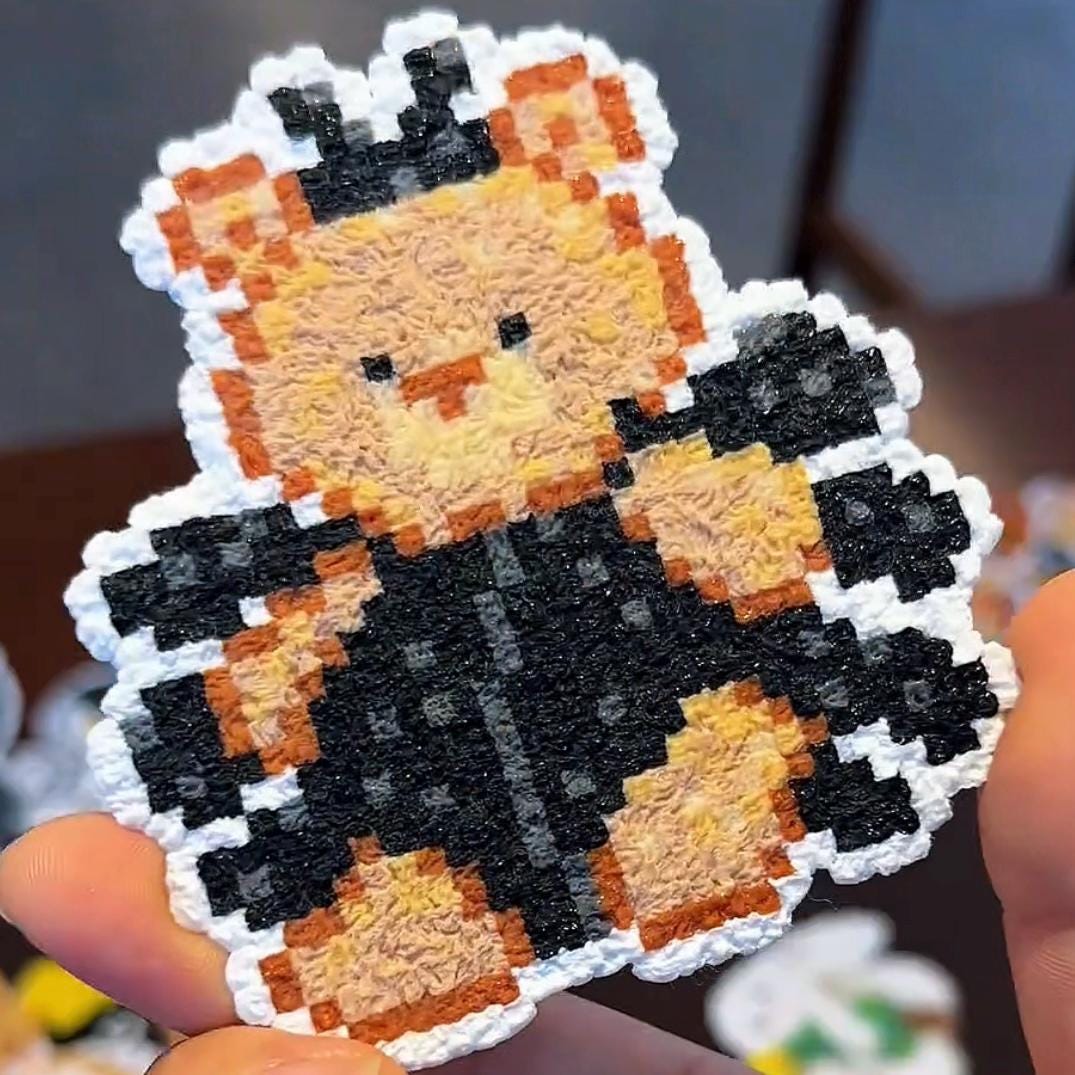 May include: A pixelated teddy bear-shaped patch wearing a black jacket and a crown. The bear is tan and orange, with black details. The patch has a white border and is held by a hand.