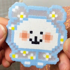 May include: A pixelated cloud-shaped charm with a white centre and a light blue outline. The charm features a smiling face with black eyes and mouth, and pink cheeks. Small white flowers with yellow centres are scattered around the edges. The charm has a