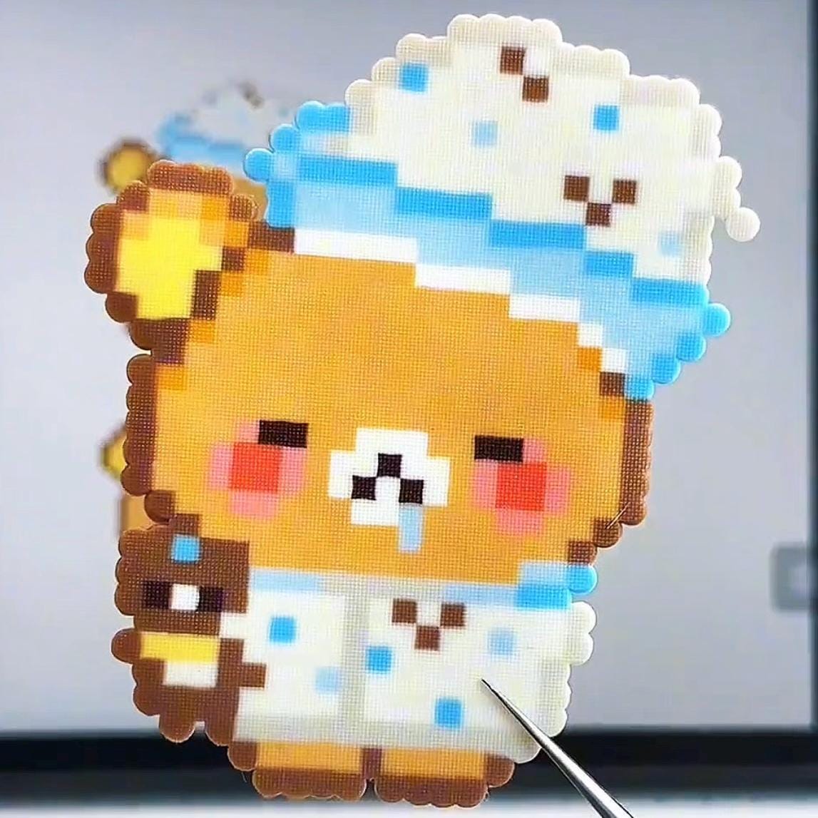 May include: Pixel art of a brown bear wearing a blue and white nightcap and pyjamas. The bear has rosy cheeks and is holding a small brown bear. The image is made of small, colourful squares.