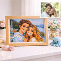 May include: A framed pixelated portrait of a smiling couple, in a wooden frame. A smaller photograph of the couple is in the upper right corner. The portrait is on a white surface, with flowers and a small blue crocheted octopus.